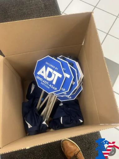 A cardboard box full of blue octagonal ADT yard signs on a tile floor.