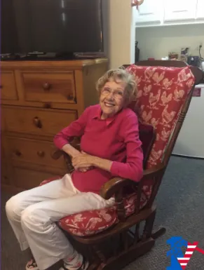 Connie, in a pink sweater, smiles from a red patterned rocking chair in her home.