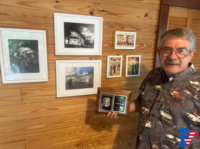 Craig holds a plaque with a silver badge beside framed firefighting photographs on a wood wall.