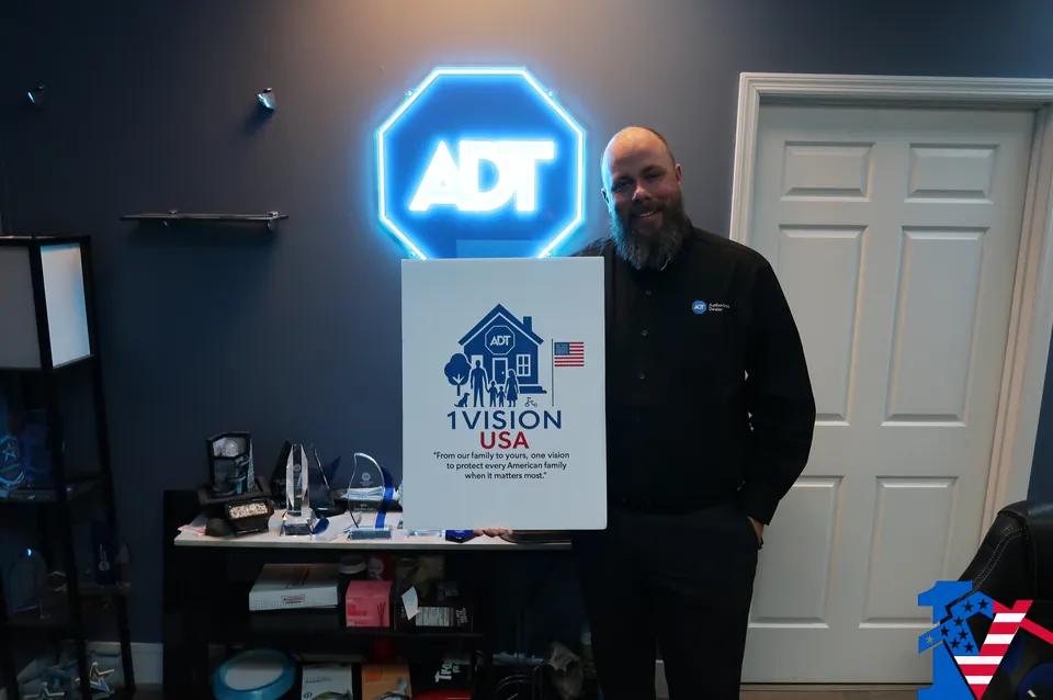 Dan stands beside a 1 Vision USA sign under a glowing blue ADT octagon light.