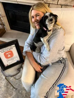 Kathleen smiles holding a small black and white dog beside a framed ASPCA flyer.