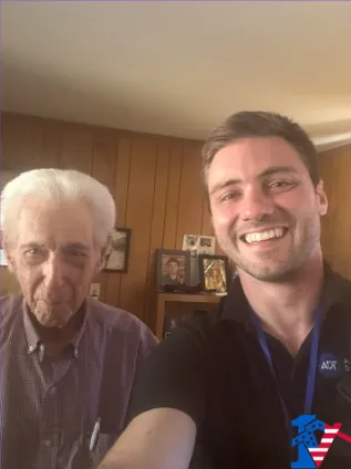 Max takes a smiling selfie with Harry, an older man, in a wood-paneled room.