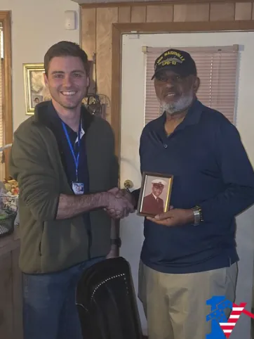 Max shakes hands with a veteran in a Navy ship cap who holds a framed portrait.