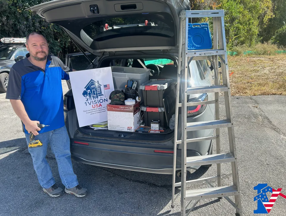 Ricki holds a drill beside an SUV trunk full of equipment bins and a 1 Vision USA sign.