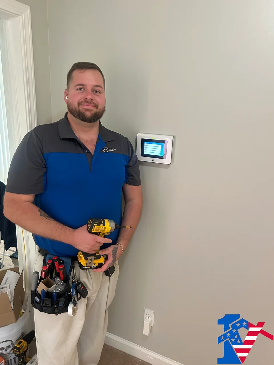 Sean, in a tool belt and holding a drill, stands beside a newly mounted security touchscreen.