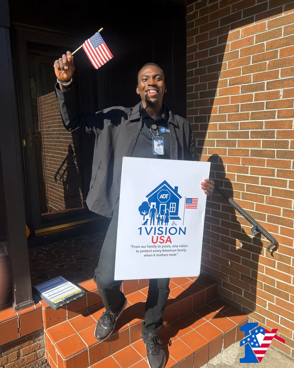Sean smiles on brick front steps, raising a small American flag and holding a 1 Vision USA sign.