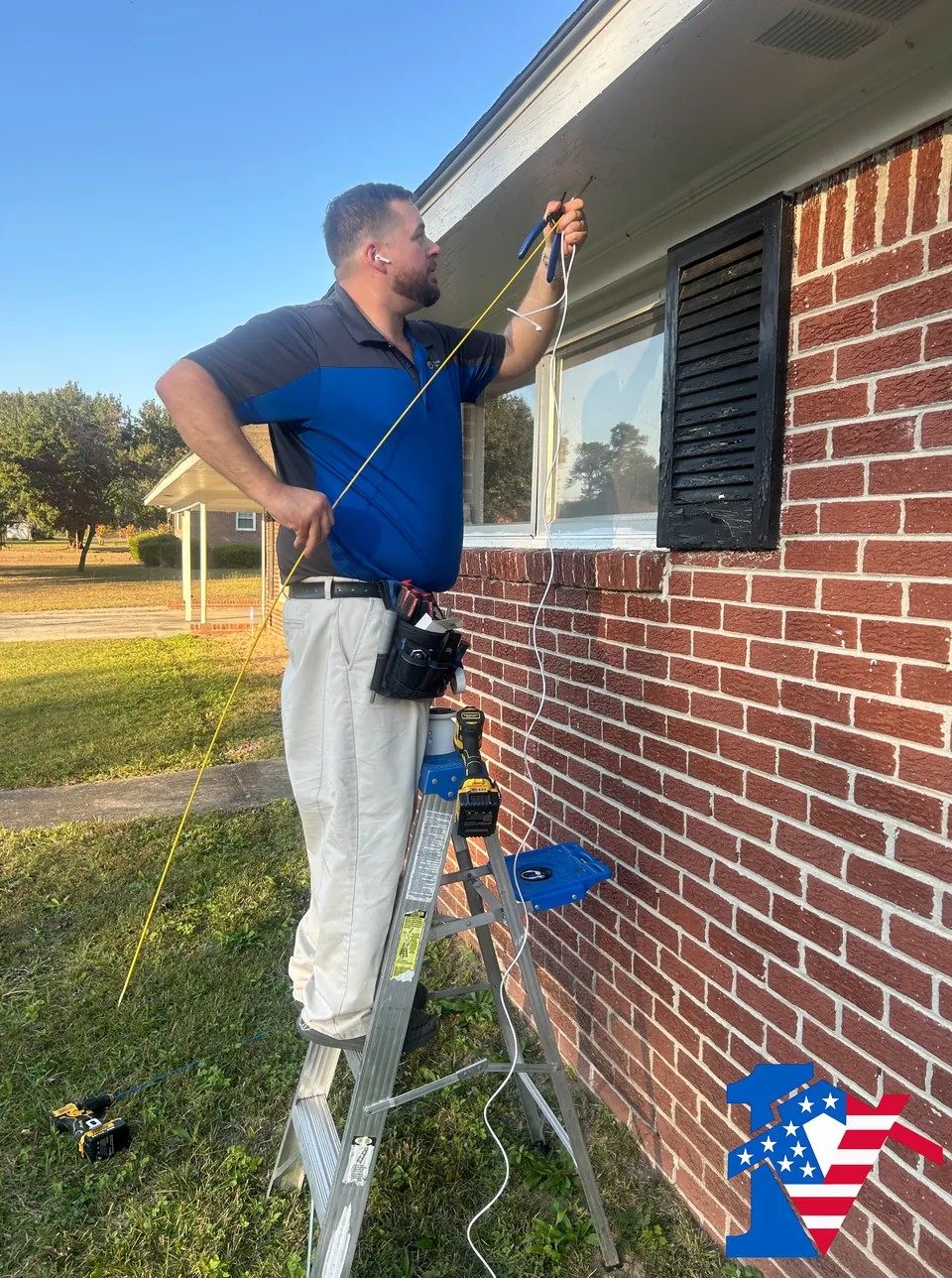 Sean, on a ladder beside a brick house, runs a cable up the wall to mount an outdoor camera.