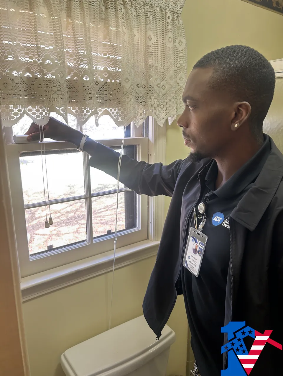 Sean fits a security sensor to a window frame behind a lace curtain.