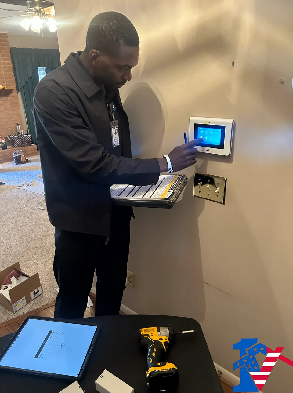 Sean taps a wall-mounted security touchscreen, a binder in hand and a drill on the table below.