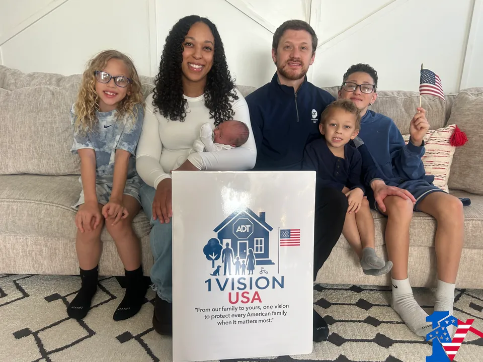 Seth's family on a couch: a woman holds a newborn, three children smile, one waves a flag, behind a 1 Vision USA sign.