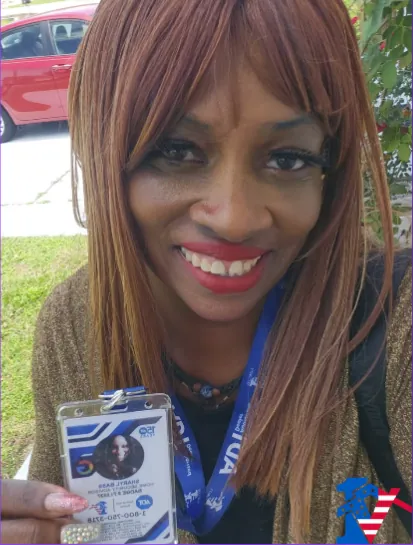 Sharyl smiles in a close selfie, holding up her 1 Vision USA ID badge on a blue lanyard.