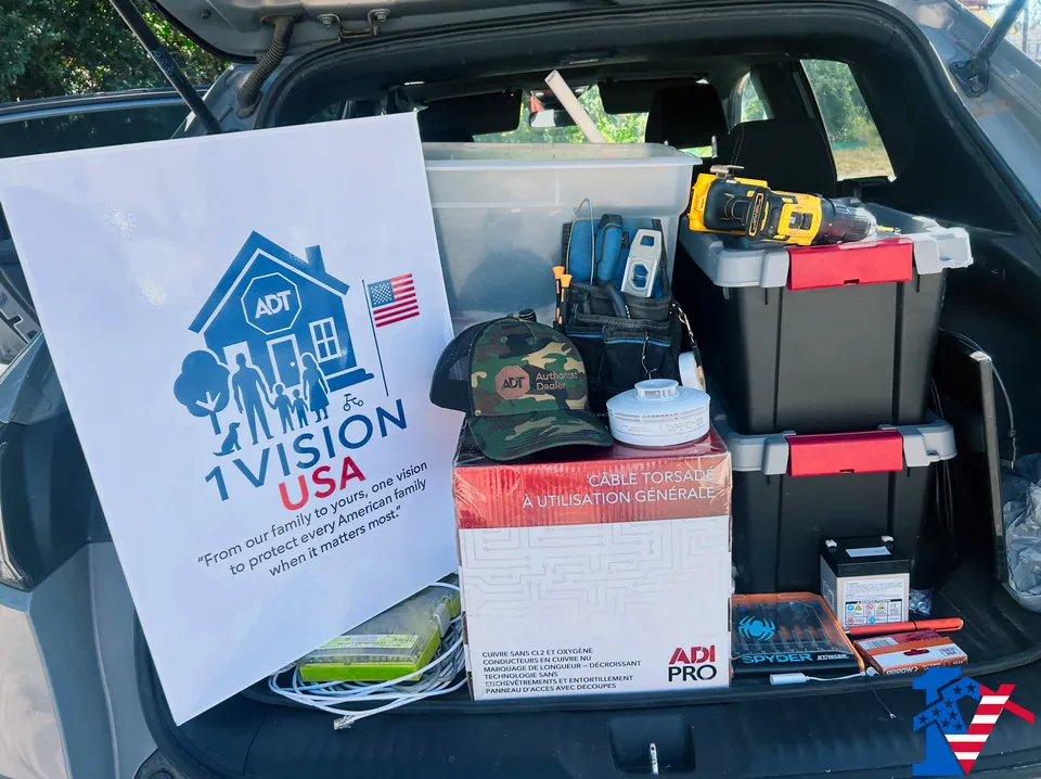 An open SUV trunk packed with equipment bins, a drill, a box of cable and a 1 Vision USA sign.