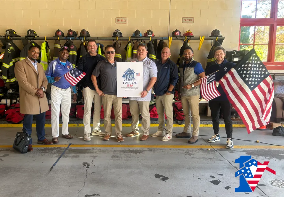 The team lines up in a fire station bay with flags and a 1 Vision USA sign, firefighter gear behind.